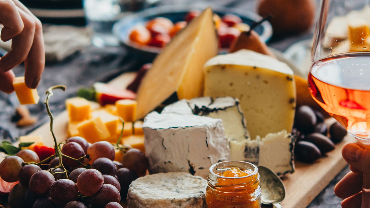A table with assorted cheeses, grapes, berries, olives, and a glass of rosé wine; hands reaching for food.