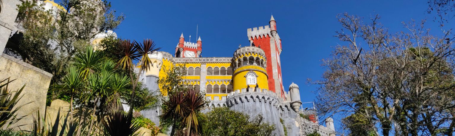 Colorful castle with red, yellow, and grey towers surrounded by lush greenery under a clear blue sky.