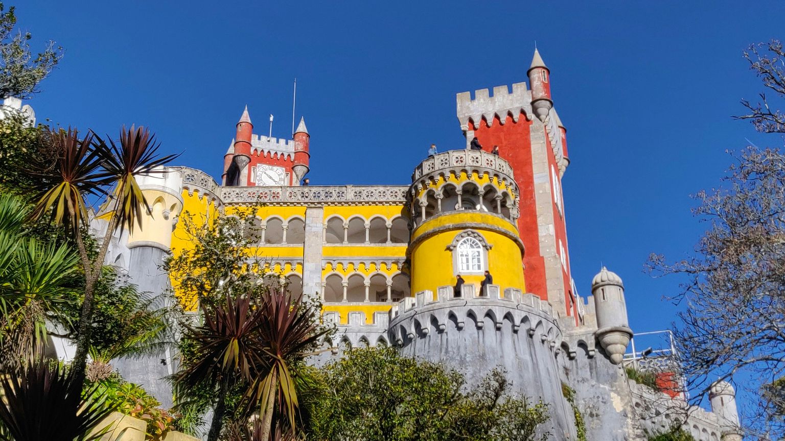 Colorful castle with red, yellow, and grey towers surrounded by lush greenery under a clear blue sky.
