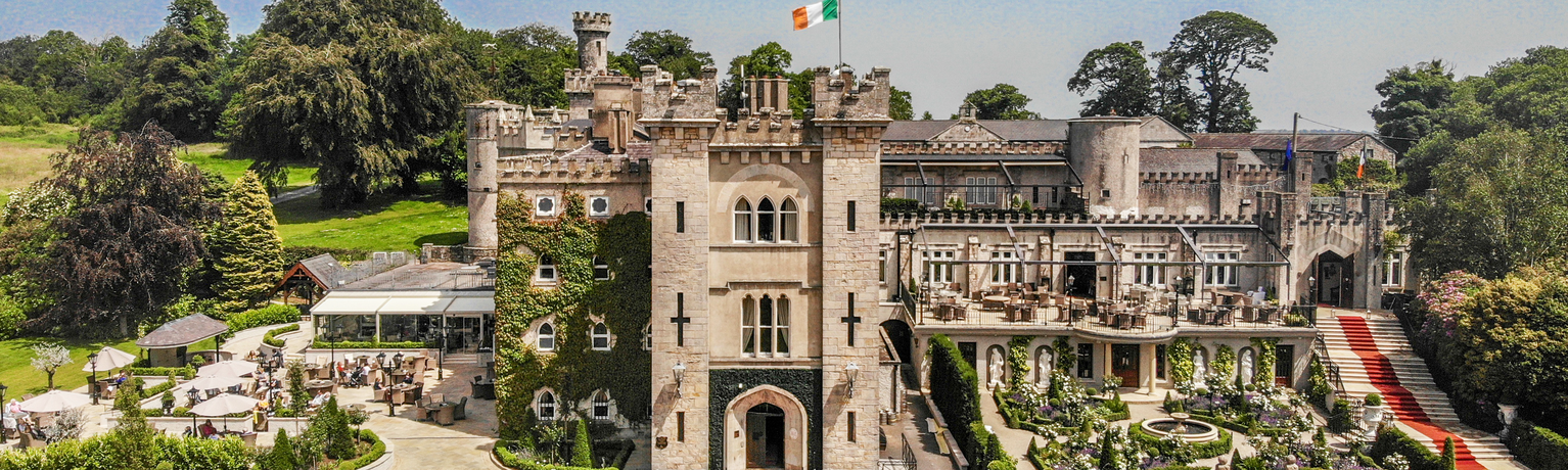 An aerial view of a stone castle with towers, an Irish flag flying on top, and an outdoor patio area with tables, umbrellas, and a red carpeted staircase.