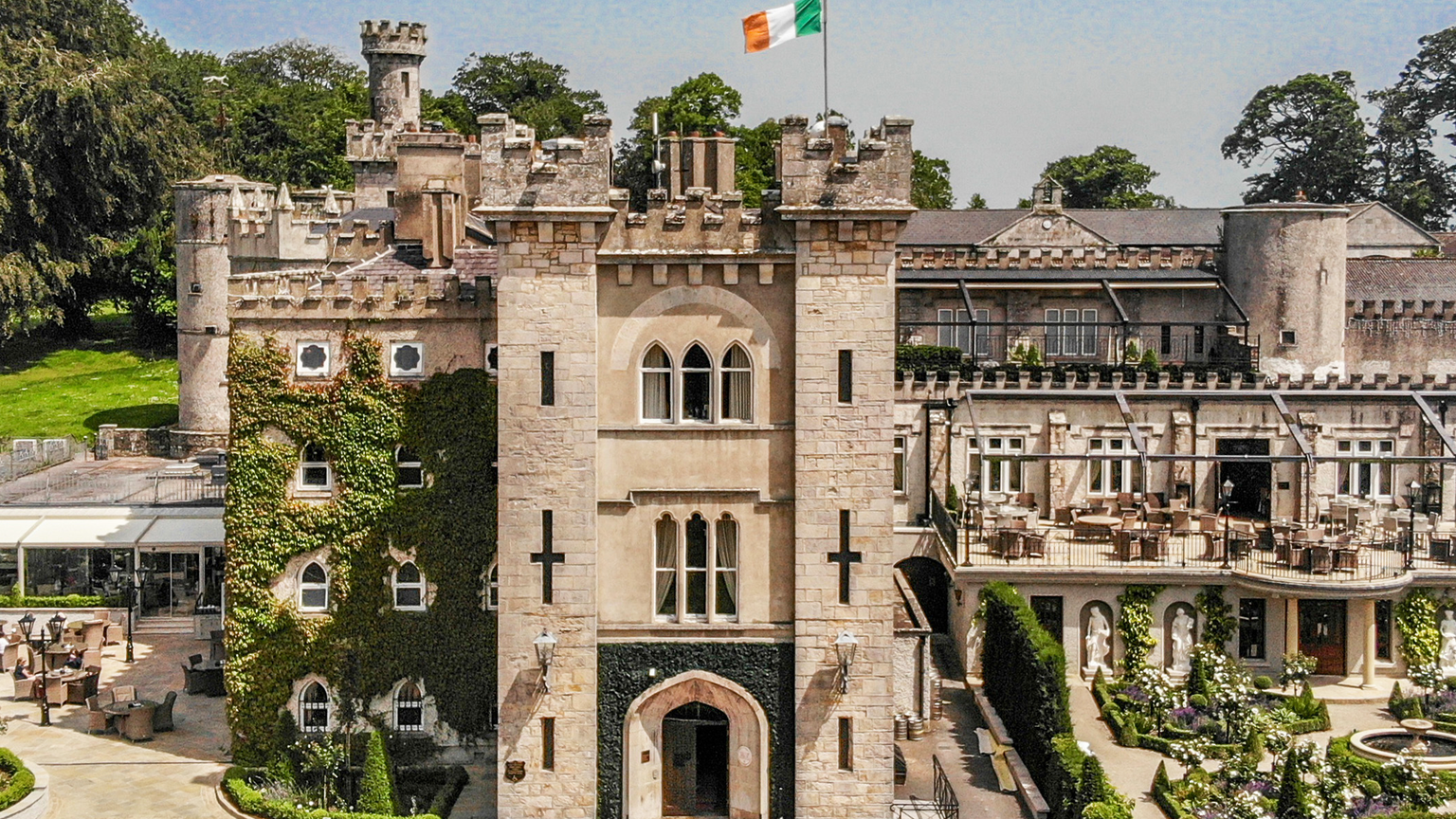 An aerial view of a stone castle with towers, an Irish flag flying on top, and an outdoor patio area with tables, umbrellas, and a red carpeted staircase.
