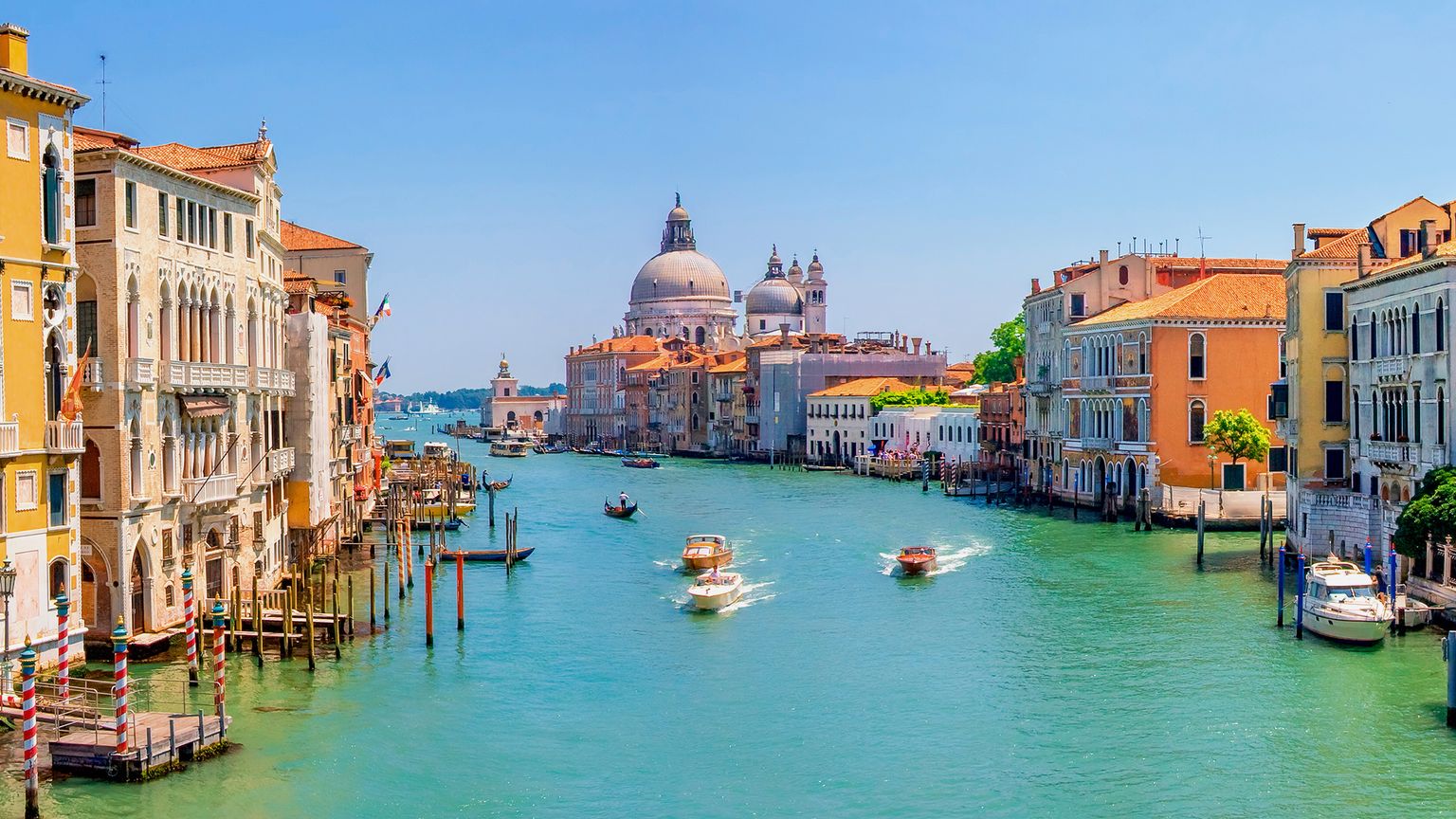 Scenic view of a Venice canal with colorful historic buildings, boats on the turquoise water, and a domed church in the background.