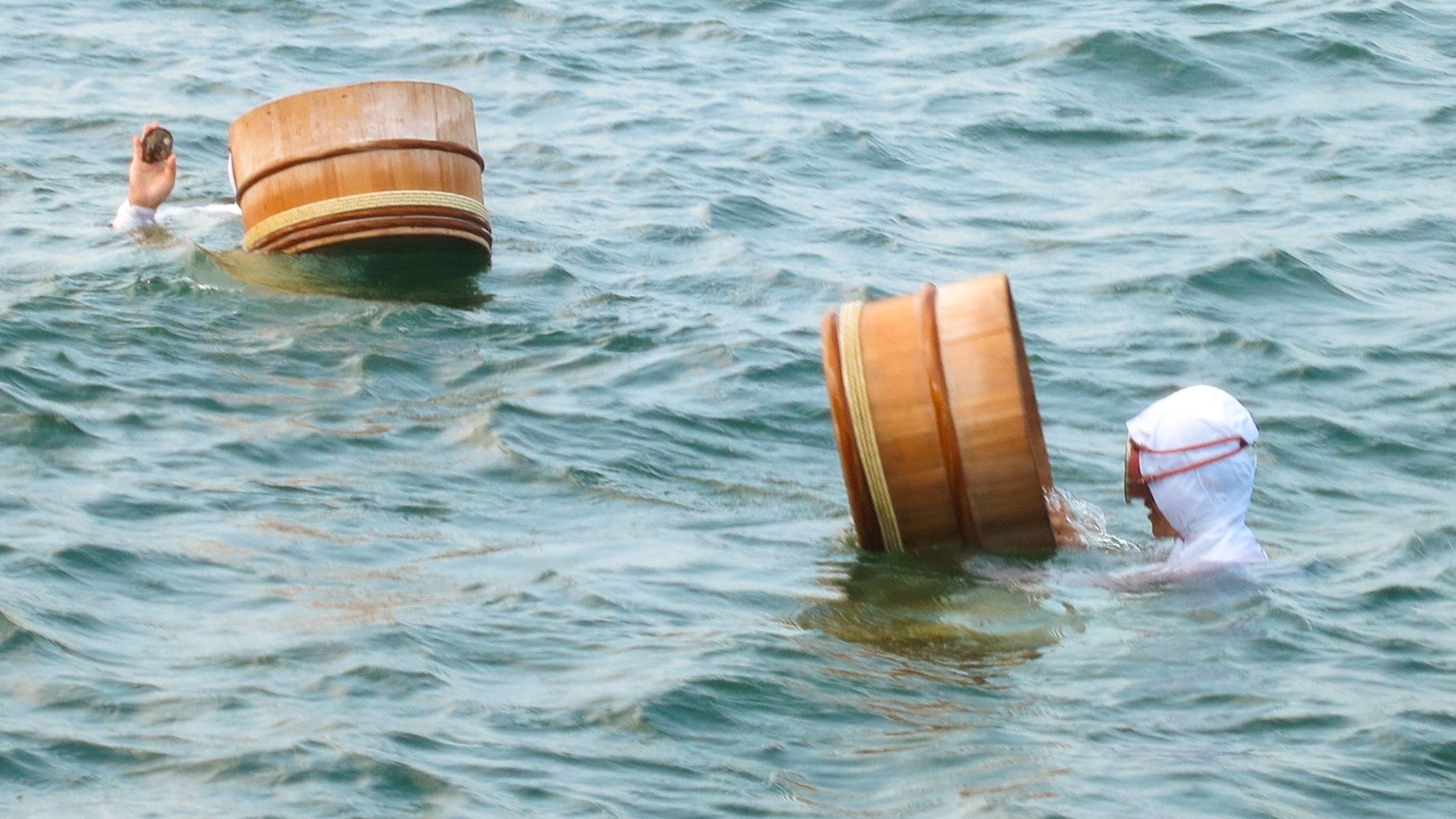 Two people in the water with traditional wooden tubs, wearing white head coverings, in a calm sea.