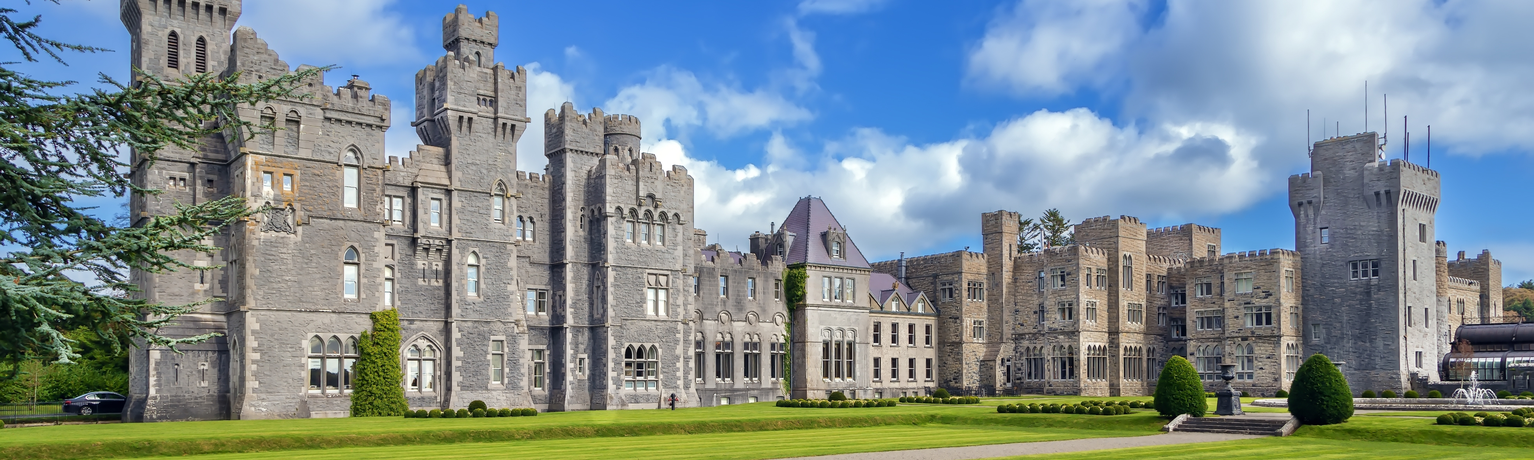 Large stone castle with multiple towers and a crenelated roofline set behind a wide lawn with manicured shrubs under a blue sky with white clouds.