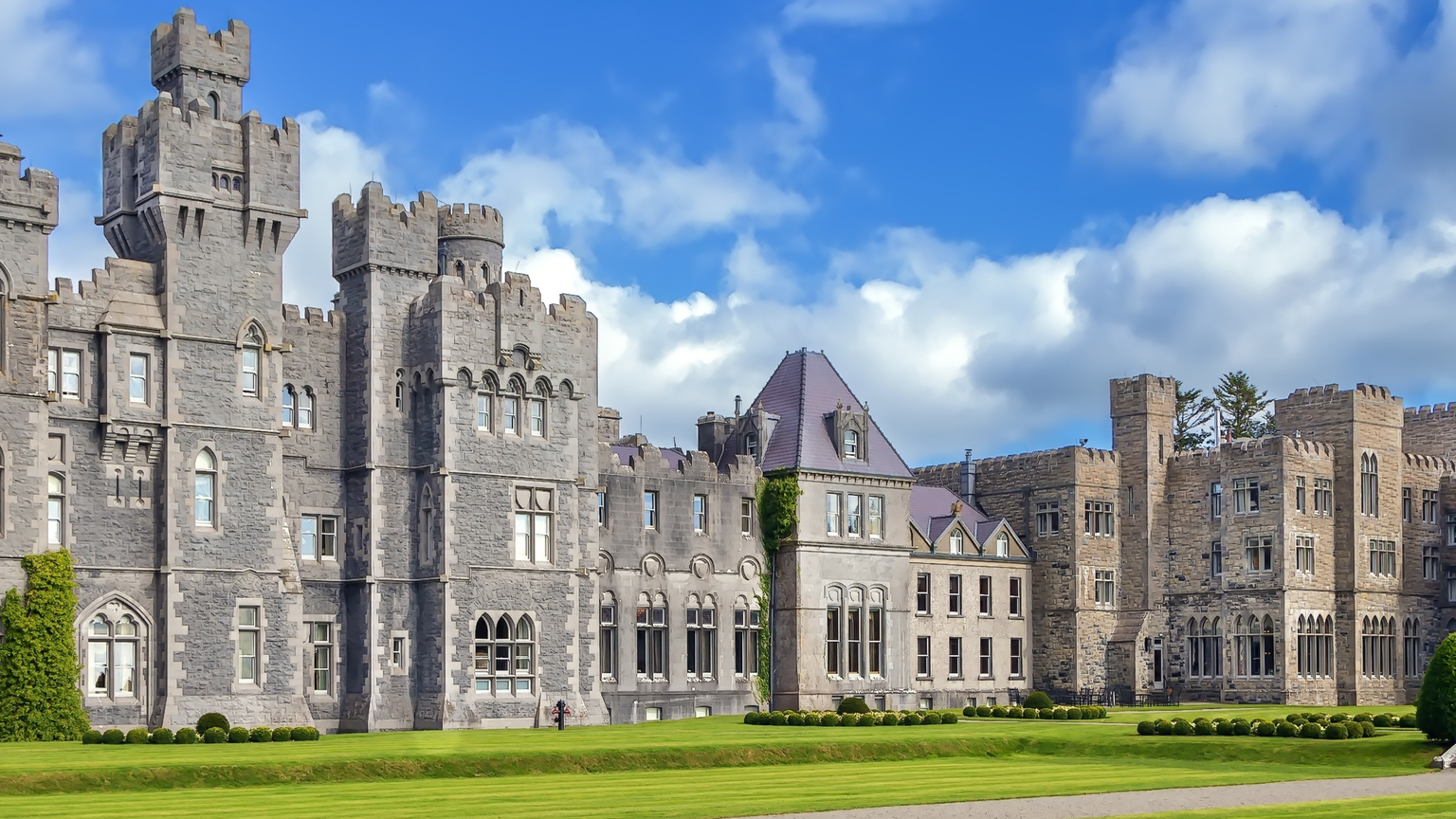 Large stone castle with multiple towers and a crenelated roofline set behind a wide lawn with manicured shrubs under a blue sky with white clouds.