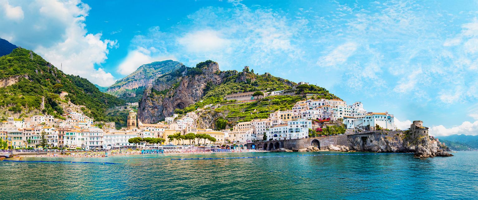 Scenic coastal town with colorful buildings nestled in lush green hills, overlooking a blue sea under a bright, cloudy sky.