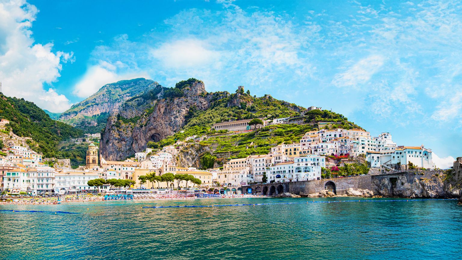 Scenic coastal town with colorful buildings nestled in lush green hills, overlooking a blue sea under a bright, cloudy sky.