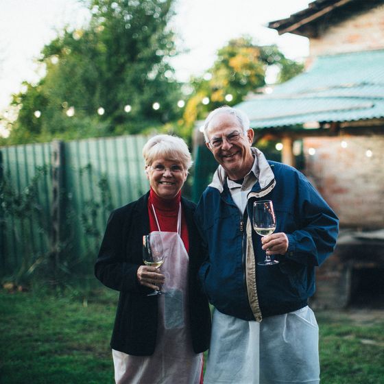 Elderly couple in aprons, smiling with wine glasses in a garden, string lights and rustic house in background.