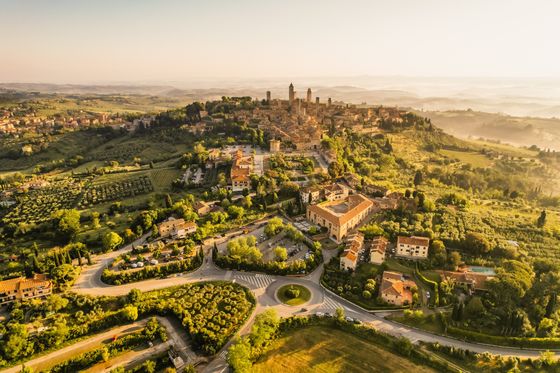 Aerial view of a Tuscan town at sunrise, surrounded by lush green fields, winding roads, and rolling hills under a golden sky.
