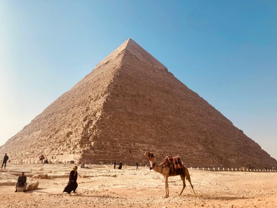 Camel and tourists near the Great Pyramid of Giza under a clear blue sky.