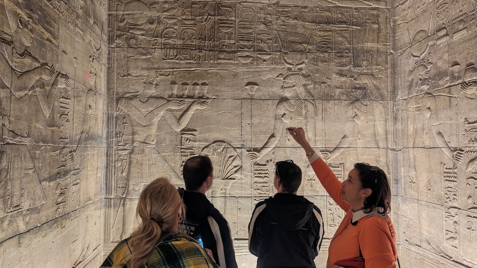 Four people observing and pointing at intricate ancient Egyptian hieroglyphs and carvings on a stone wall inside a dimly lit room.