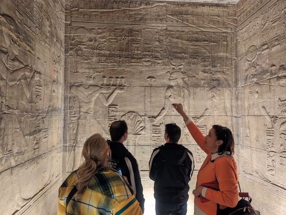 Four people observing and pointing at intricate ancient Egyptian hieroglyphs and carvings on a stone wall inside a dimly lit room.