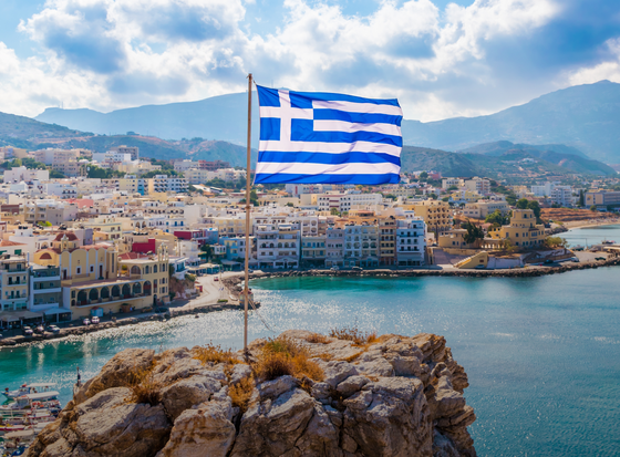 Greek flag on a rocky hill overlooking a coastal town with colorful buildings and a blue sea under a partly cloudy sky.