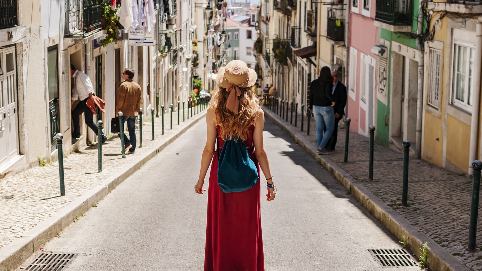 A woman in a red dress and straw hat stands facing downhill on a narrow, colorful street, with people walking in the background.