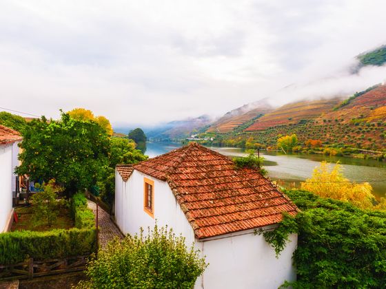White houses with red-tiled roofs, surrounded by lush greenery, overlooking a river and terraced hills on a cloudy day.