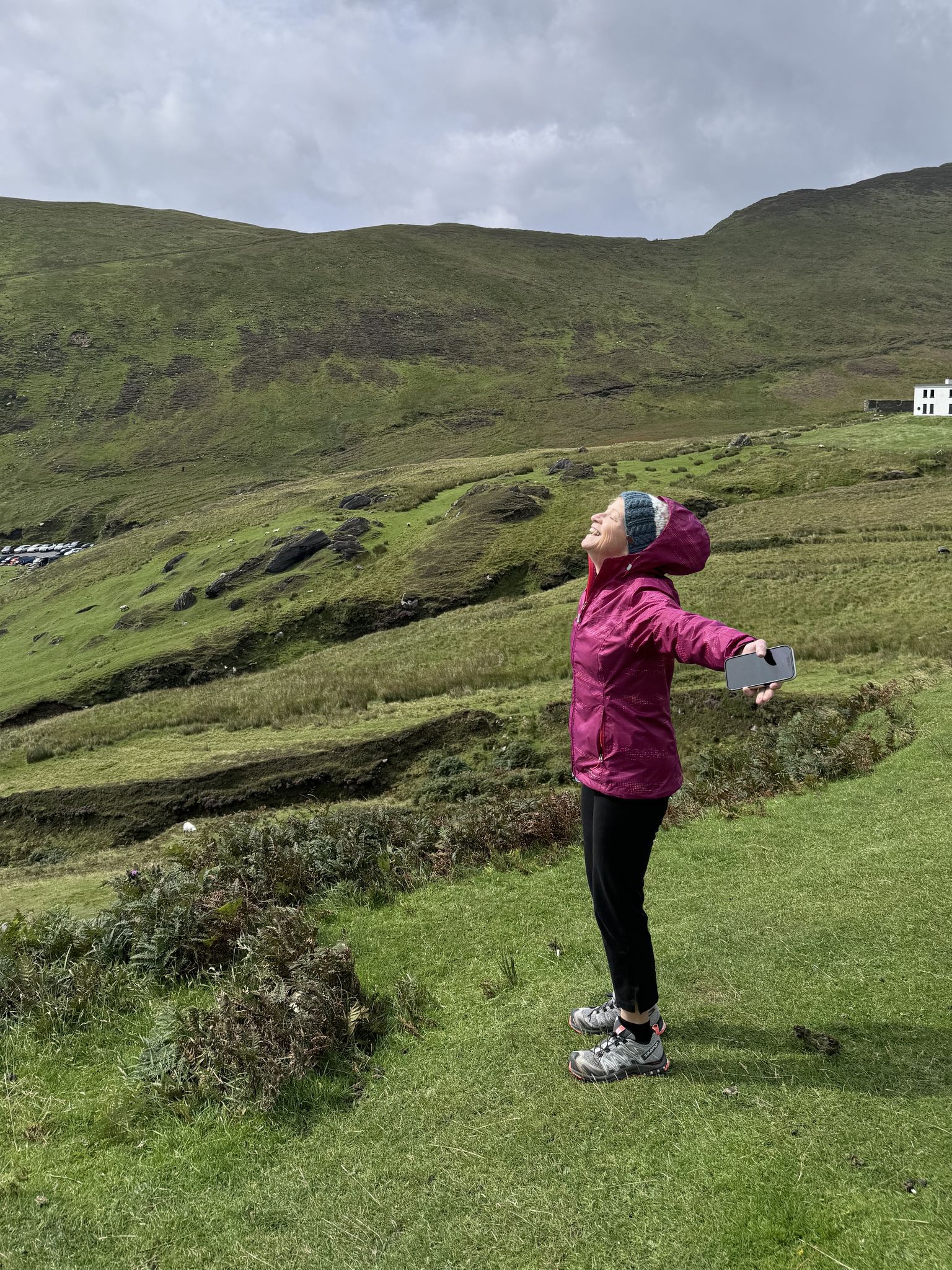 Person in a pink jacket and blue hat enjoying the outdoors, stands on a grassy hill with arms outstretched, surrounded by rolling green hills.