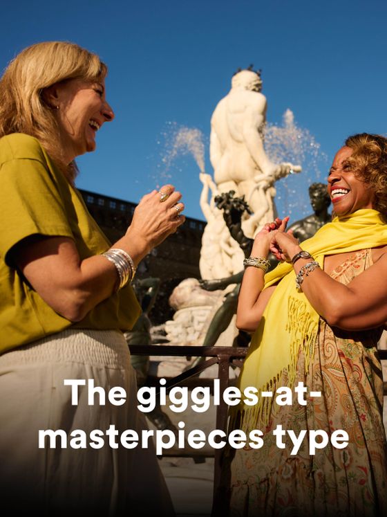 Two women laughing in front of a fountain sculpture under a clear blue sky, with text overlay: "The giggles-at-masterpieces type."