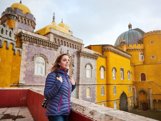 A woman in a blue jacket stands smiling on a terrace, with the vibrant yellow and purple walls of the Pena Palace in Sintra in the background.