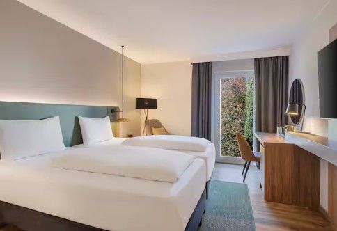 Modern hotel room with two neatly made beds, a wooden desk with a lamp, a chair, and a large window with curtains overlooking greenery.