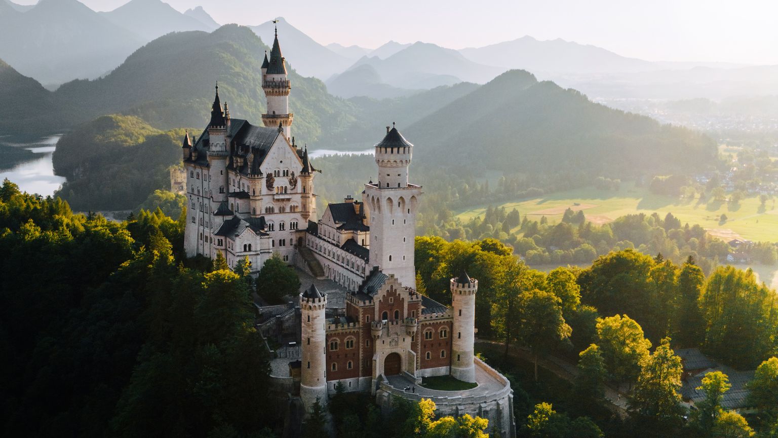 Aerial view of Neuschwanstein Castle nestled among lush green forests, with a backdrop of mountainous landscape under a clear sky.