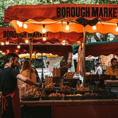 Vibrant outdoor market scene at Borough Market, with a vendor serving food under red canopies, surrounded by stalls and people.