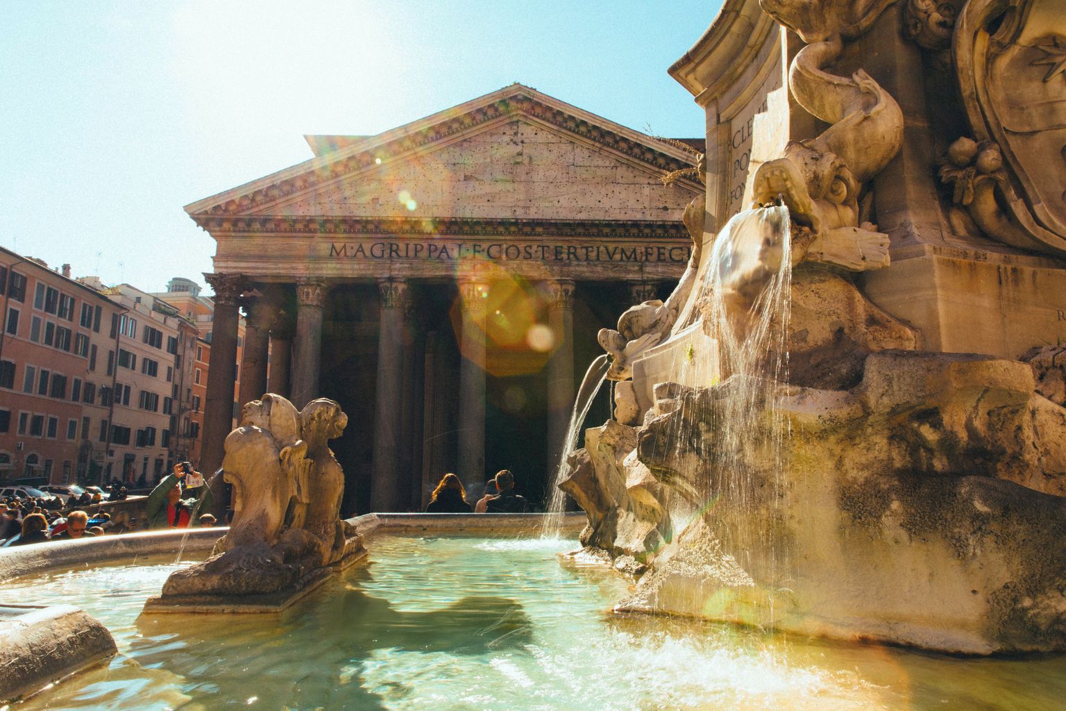 Fountain in front of the Pantheon in Rome under a clear sky, with sunlight creating lens flares.