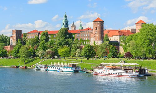 View of Wawel Castle by the Vistula River with tour boats docked along the shore, set against a backdrop of trees and a blue sky.