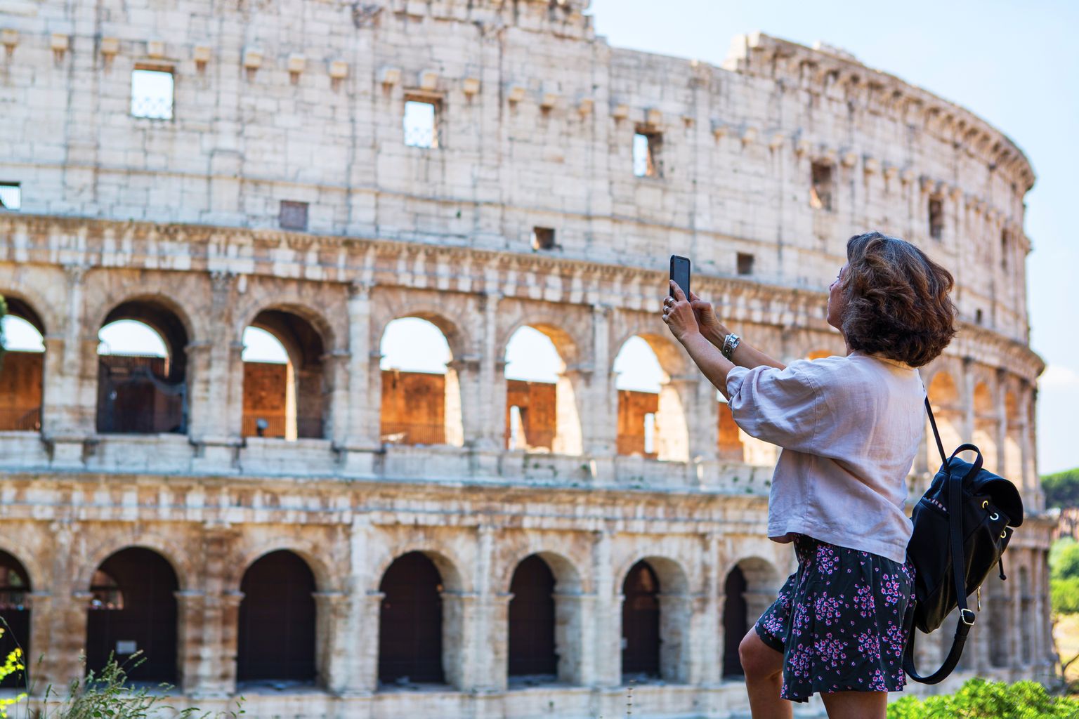 Woman taking a photo of the Colosseum in Rome, wearing a white shirt, floral skirt, and backpack, with a clear blue sky backdrop.