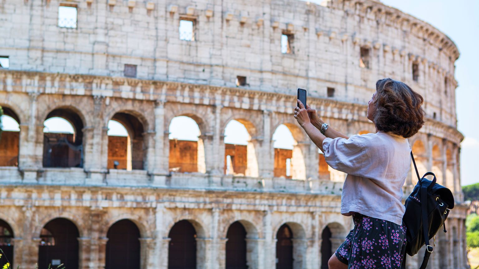 Woman taking a photo of the Colosseum in Rome, wearing a white shirt, floral skirt, and backpack, with a clear blue sky backdrop.