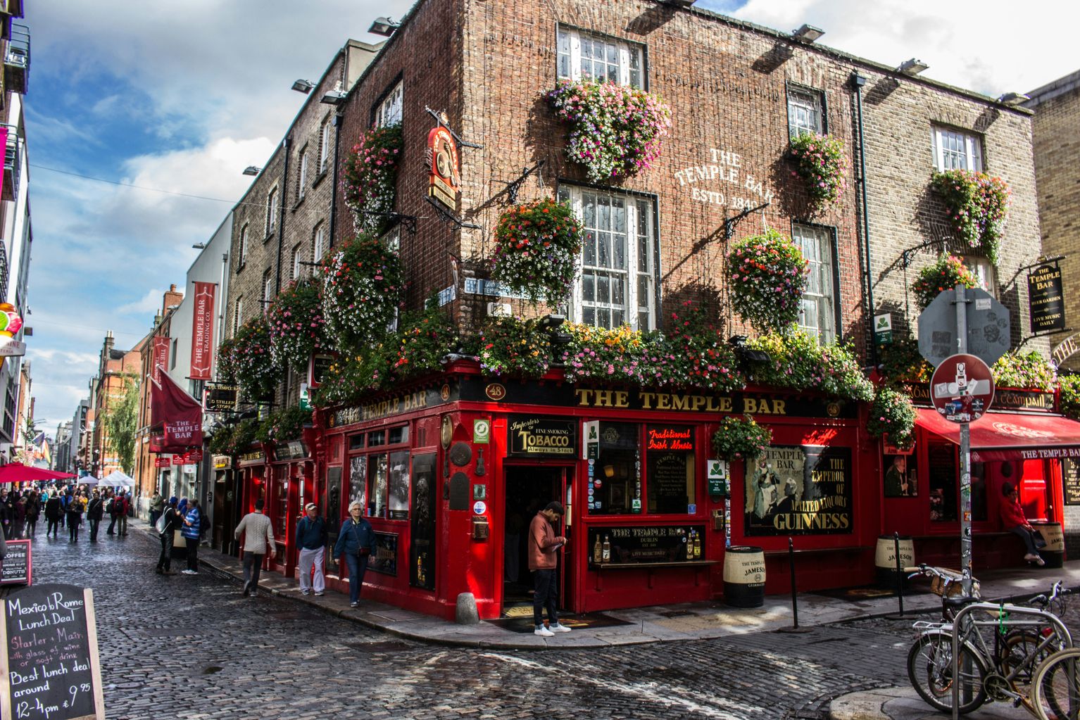 The Temple Bar pub in Dublin, a brick building with hanging flower baskets, is situated on a cobblestone street corner. People are walking on the sidewalk, and a person is standing in the doorway of the red-painted storefront, which features signs reading "The Temple Bar" and "The Emperor of Malted Liquors Guinness."