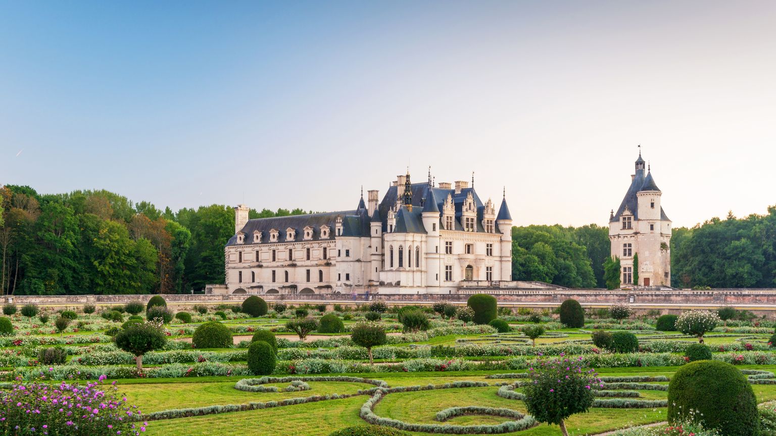 Elegant château framed by lush gardens and trees, set against a clear blue sky at dusk.
