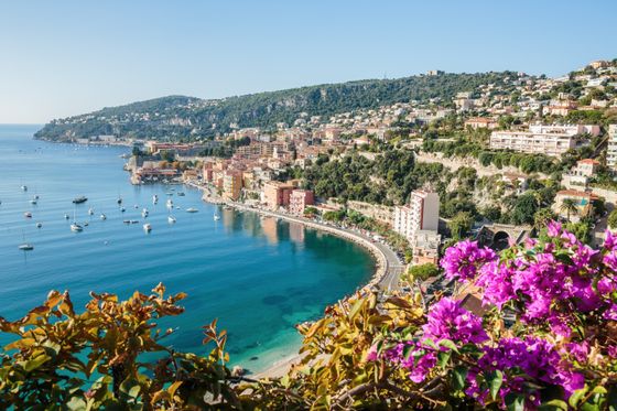 Scenic coastal view of Villefranche-sur-Mer, France, with colorful buildings, boats on blue water, and vibrant flowers in the foreground.