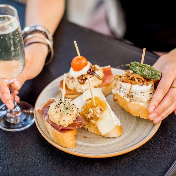 A person holds a glass of sparkling wine and a plate with assorted gourmet tapas, including cheese, prosciutto, cherry tomato, and peppers.