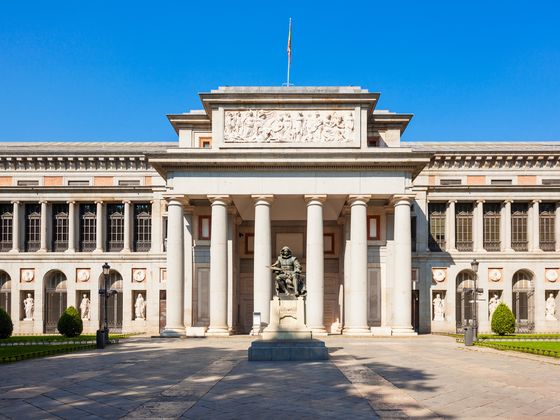 Front view of the Prado Museum in Madrid, featuring neoclassical architecture with large columns and a statue in the foreground under a clear blue sky.