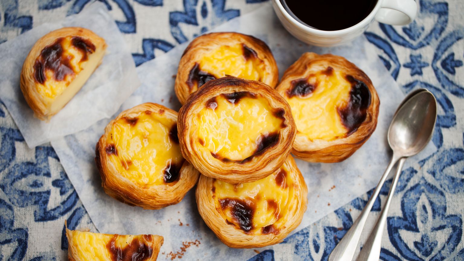A plate of Portuguese custard tarts on a blue patterned tablecloth, next to a cup of coffee and two spoons.