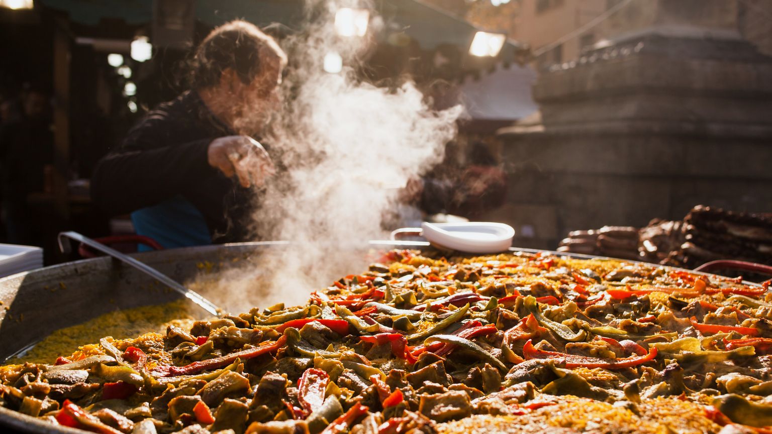 A large pan of steaming paella with colorful vegetables and meat is being served at an outdoor market stall.
