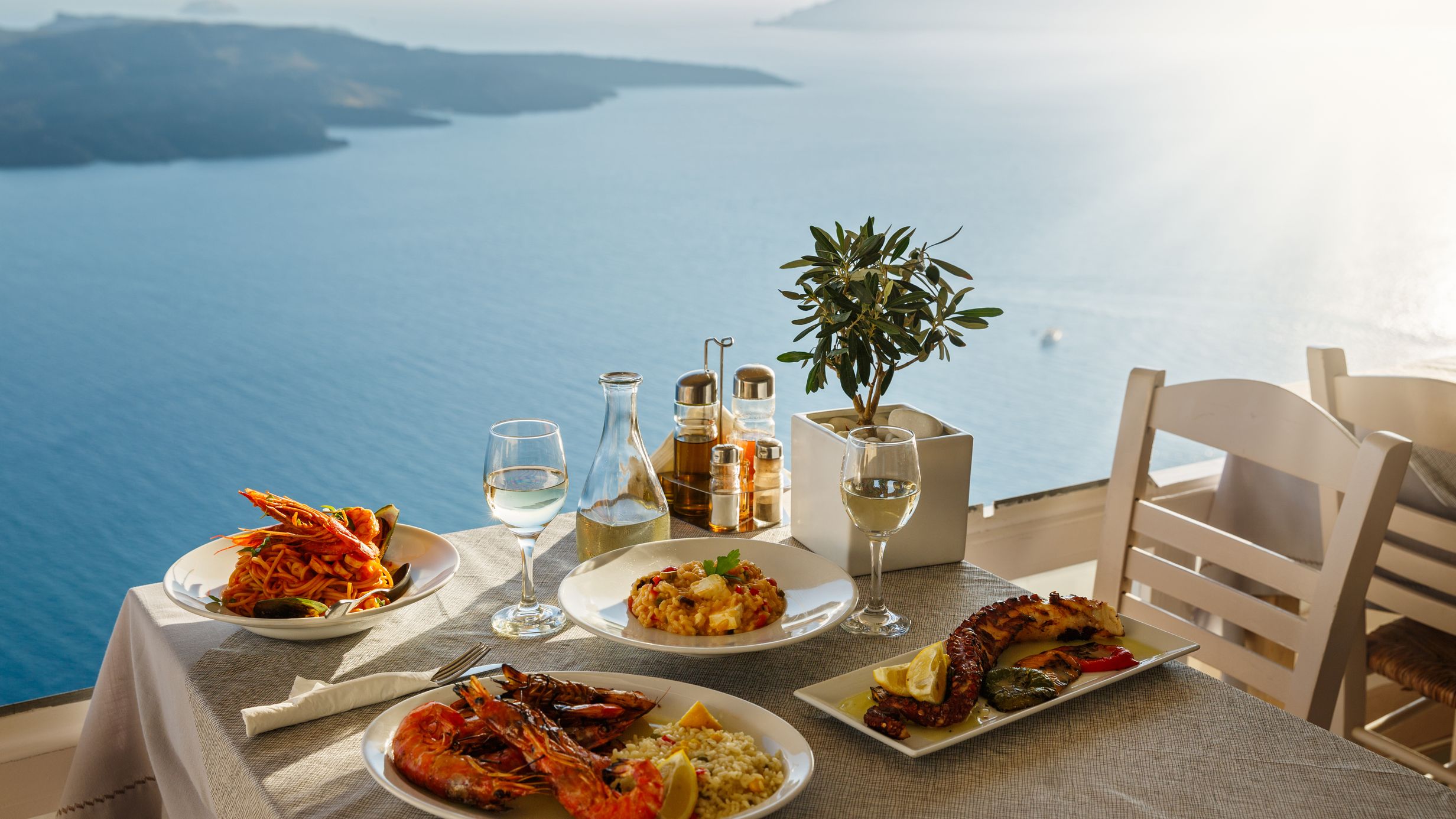 A seaside dining scene with seafood dishes, two glasses of wine, and an olive plant, overlooking a sunny ocean view.