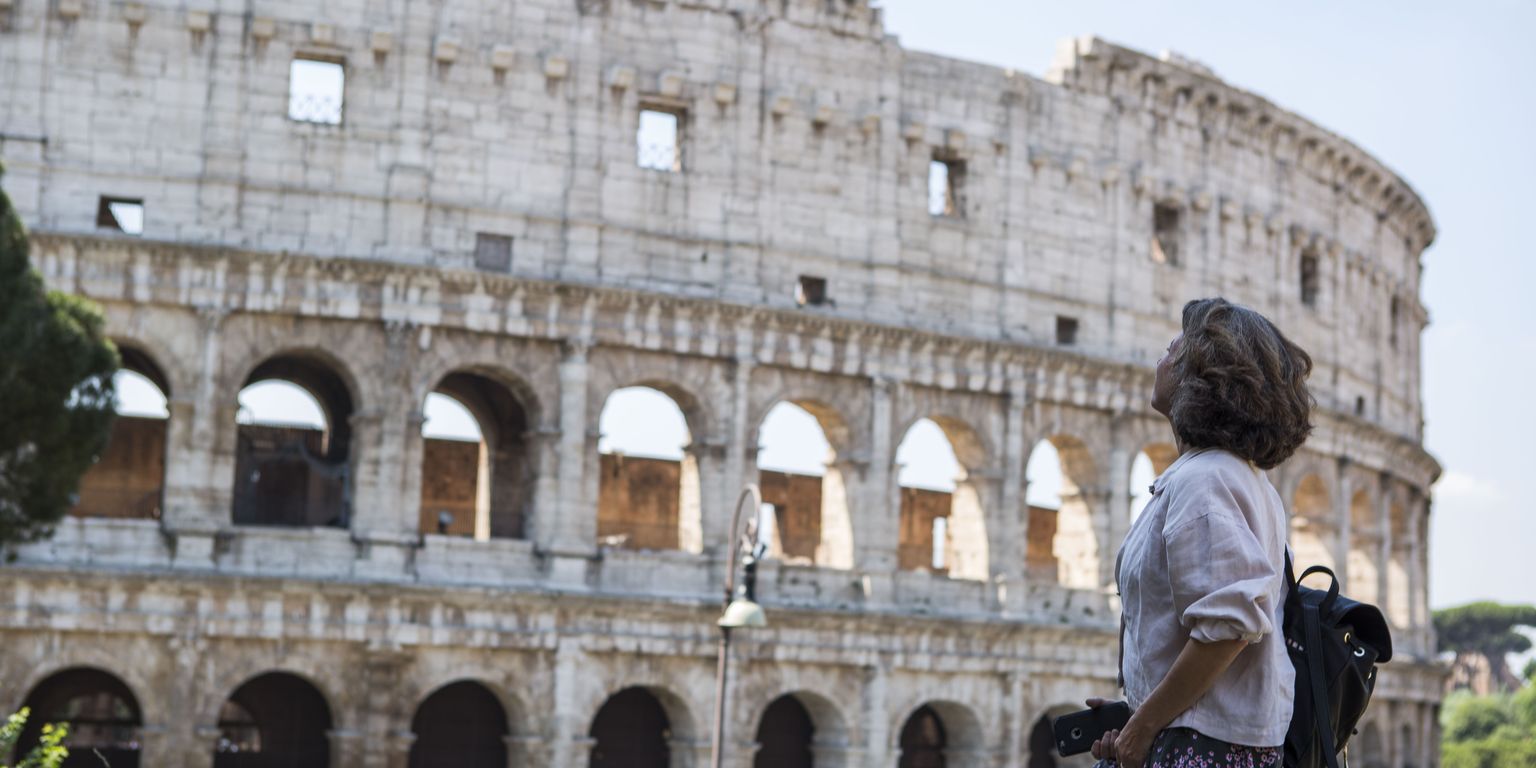 A person stands, looking up at the Colosseum in Rome, wearing casual clothing and holding a camera. The sky is clear.