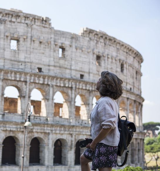 A person stands, looking up at the Colosseum in Rome, wearing casual clothing and holding a camera. The sky is clear.