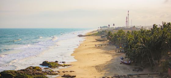 A scenic beach with gentle waves, scattered people, palm trees, and boats under a hazy sky with communication towers in the background.