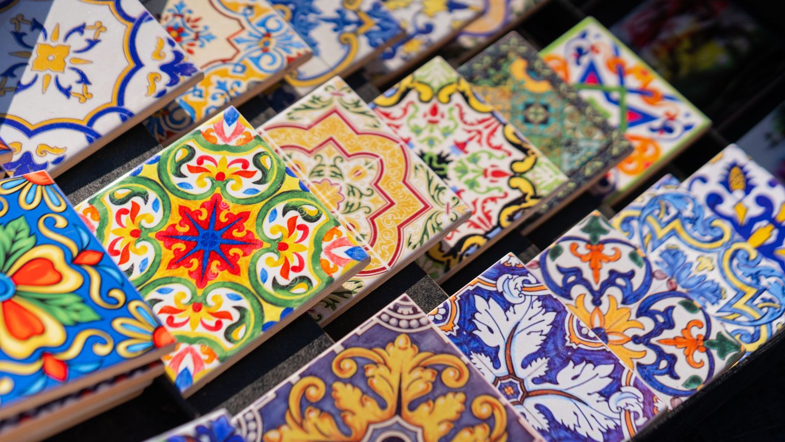 Colorful ceramic tiles, featuring intricate and varied floral and geometric patterns, arranged in rows and overlapping slightly.