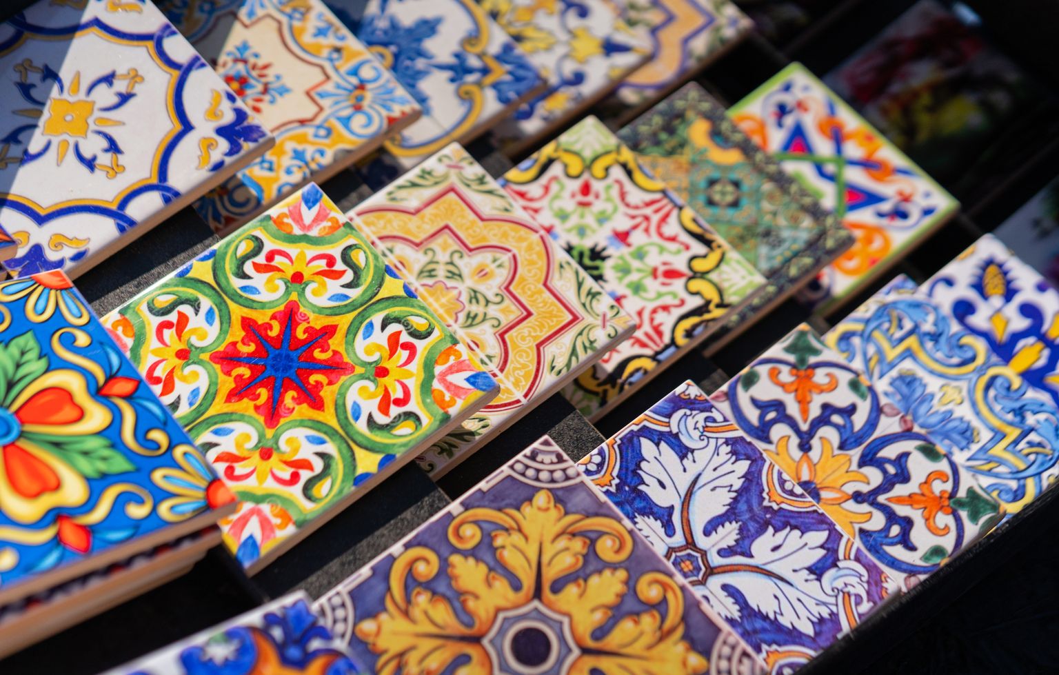 Colorful ceramic tiles, featuring intricate and varied floral and geometric patterns, arranged in rows and overlapping slightly.