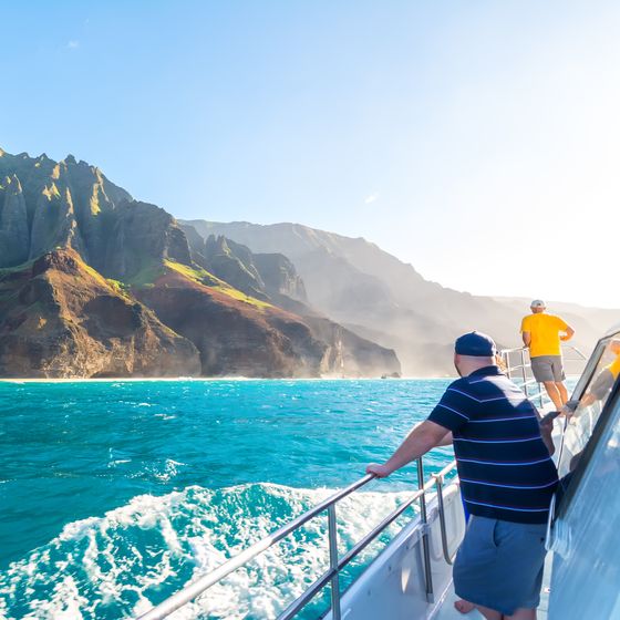 People on a boat admire the rugged, green cliffs of a coastline under a clear blue sky, with vibrant turquoise ocean water below.