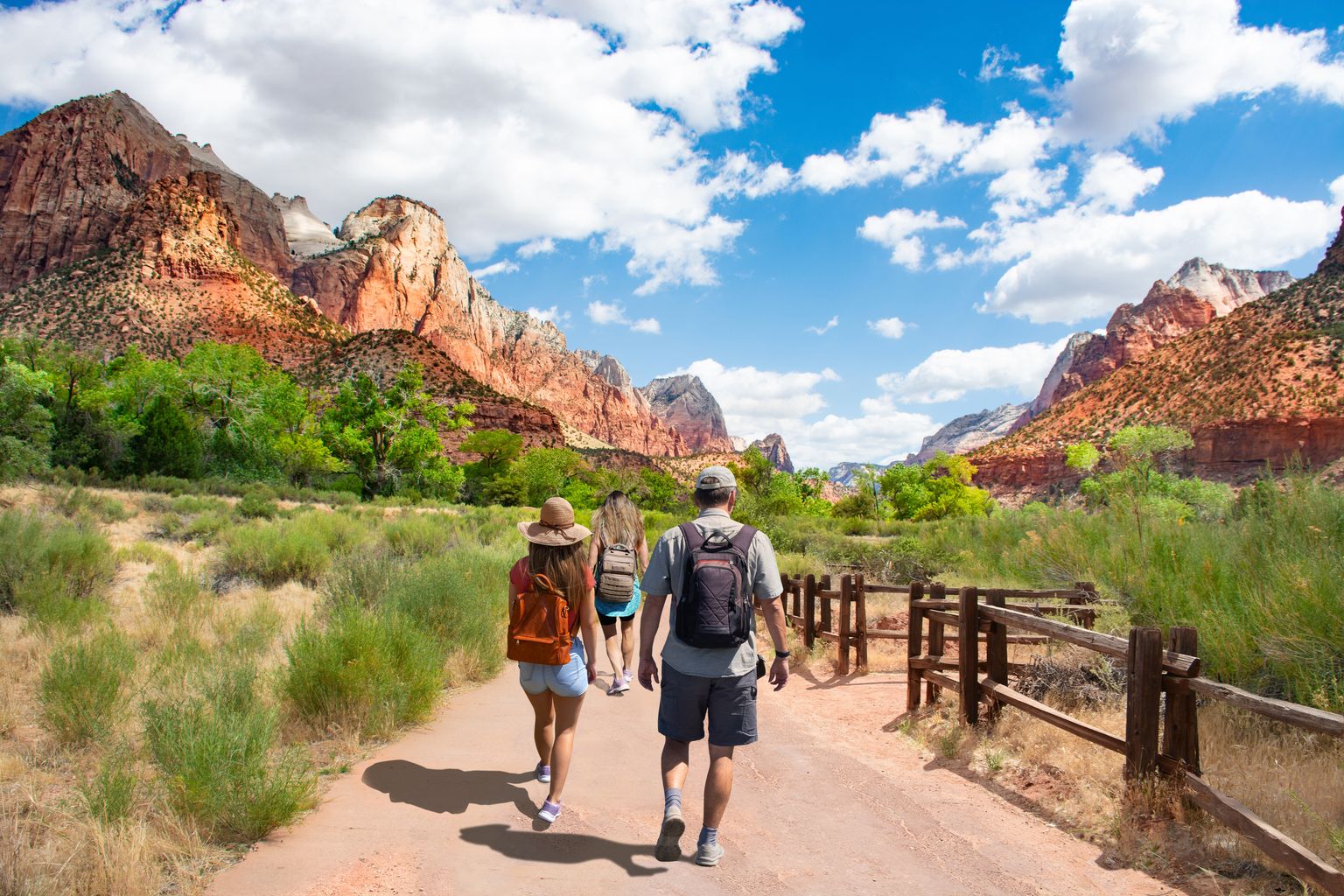 Hikers walk on a trail surrounded by red rock cliffs and greenery under a bright blue sky with scattered clouds.