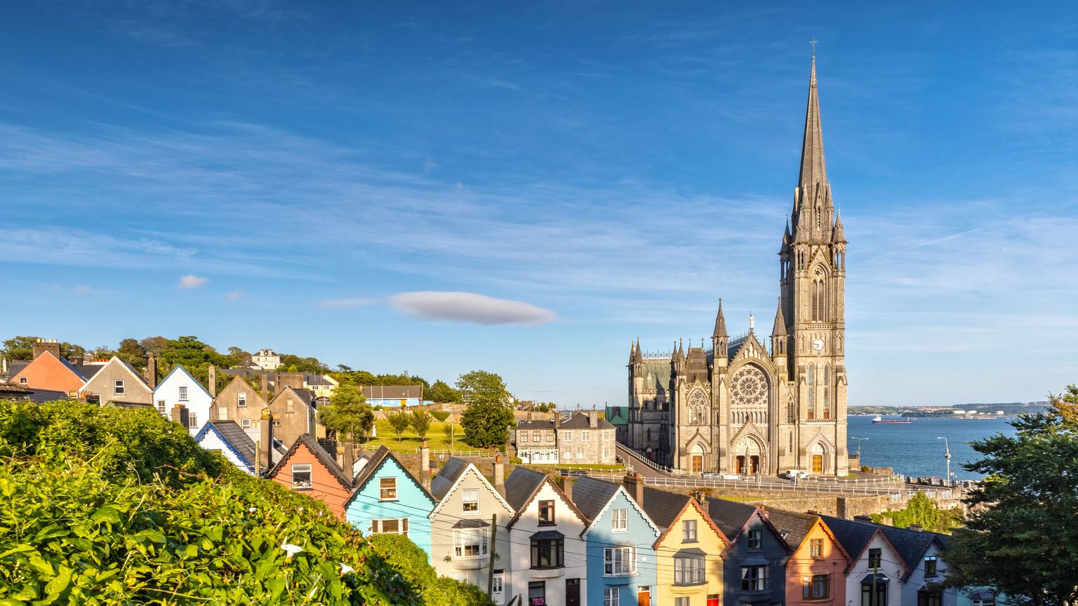 Colorful houses line a street below the towering spire of a Gothic-style cathedral, set against a backdrop of blue sky and distant water.