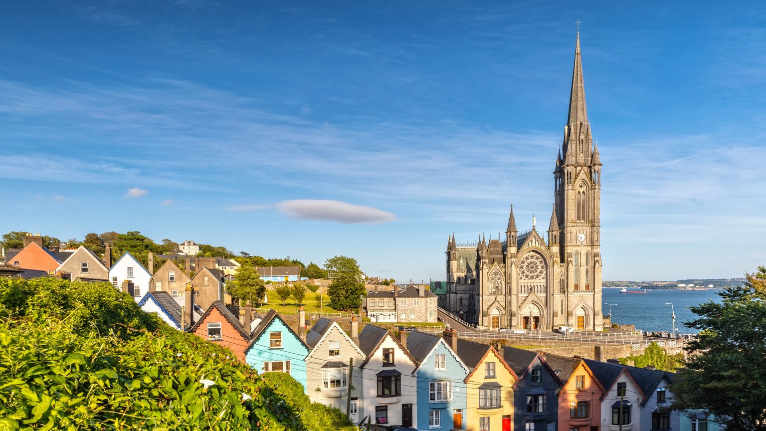 Colorful houses line a street below the towering spire of a Gothic-style cathedral, set against a backdrop of blue sky and distant water.