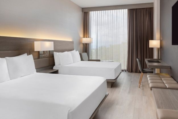 Modern hotel room with two neatly made double beds, a desk, seating area, and large window with sheer curtains. Neutral tones create a calming atmosphere.