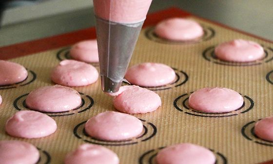 Piping pink macaron batter onto a baking mat with pre-printed circles, aligning for even size and shape.