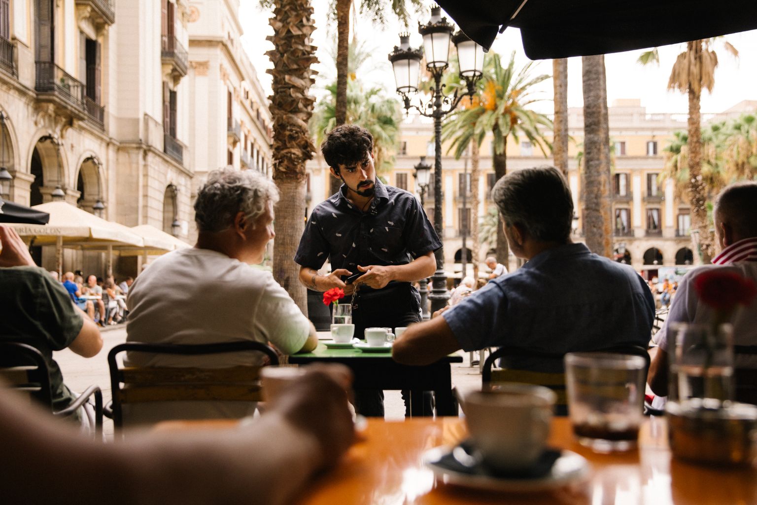 A waiter takes an order from customers seated at an outdoor café surrounded by palm trees and historic architecture.
