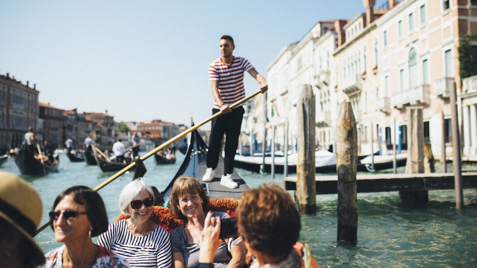 A gondolier navigates a group of smiling tourists through Venice's canals on a sunny day, with colorful buildings lining the water.